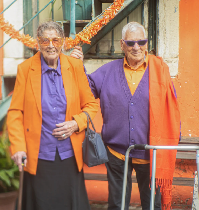 An elderly couple wearing coordinating outfits that are orange and purple. He has a walker and she has a cane.