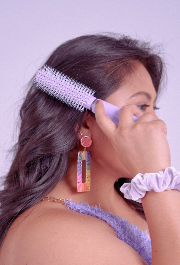 Side view of a woman brishing her hair with a purple hairbrush. She is wearing a purple shirt, a purple scrunchie on her wrist in front of a purple background