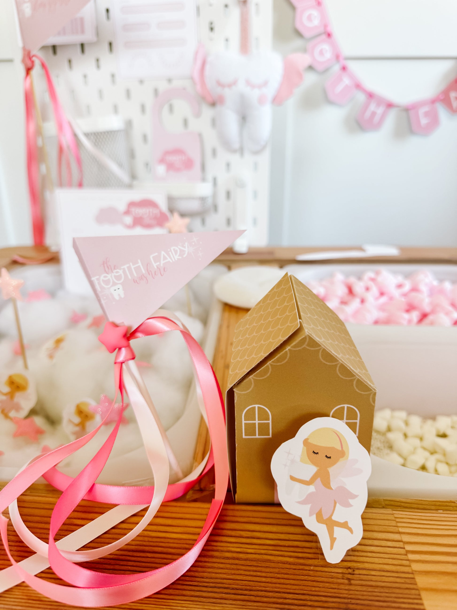 Pink ribbon gift bag and small cardboard tooth fairy house with angel cutout on table