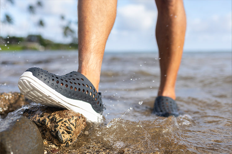 Men’s black perforated slip-on water shoes on rocky surface with cushioned sole