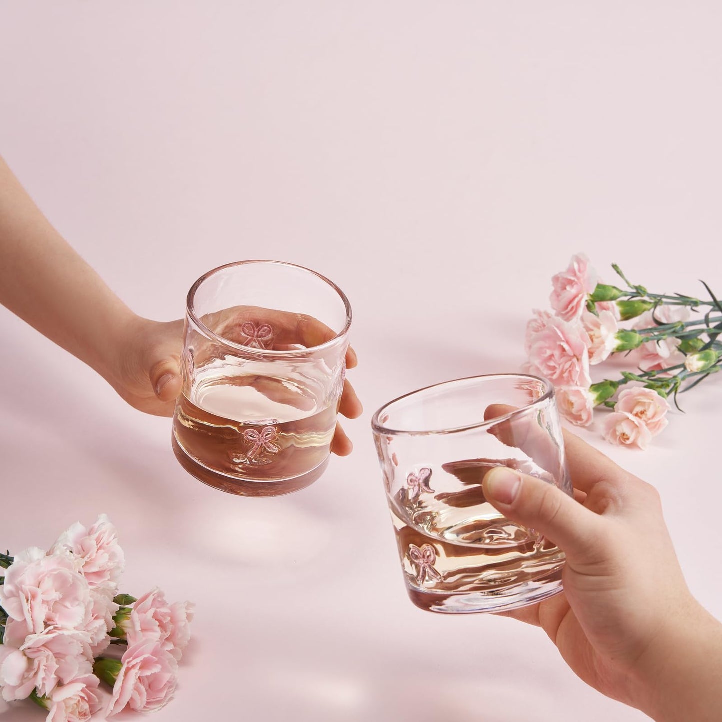 Two clear acrylic tumblers with pink bowtie motifs clinking, golden drink inside