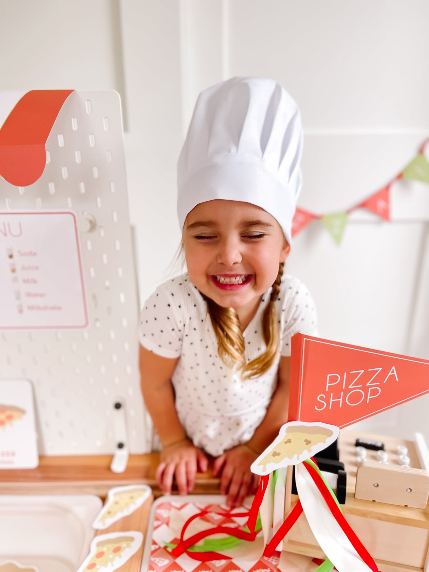 Pizza shop play kit with chef-hat child, pizza shop sign, and pretend toppings on tray