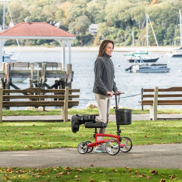 Red knee scooter in use with person standing; black basket and knee rest visible
