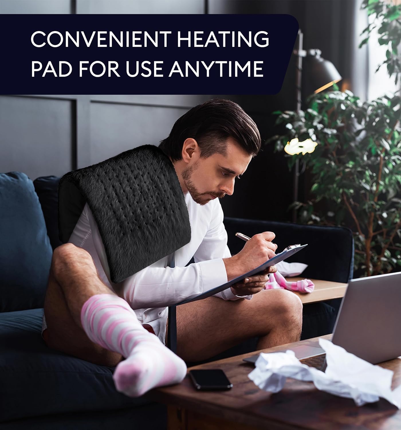 Black heating pad with controller in hand while a person reclines on a sofa