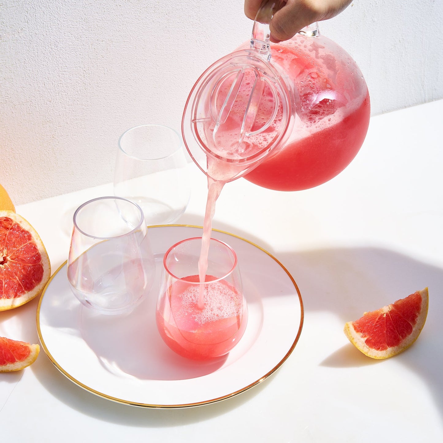 Clear acrylic pitcher pouring pink drink into glasses, with grapefruit slices and citrus