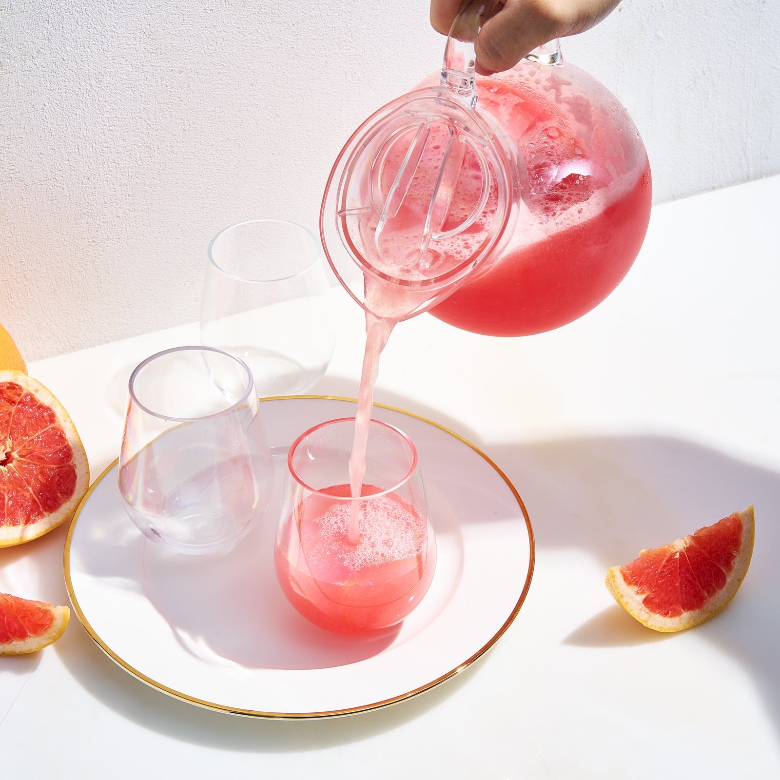 Clear acrylic pitcher pouring pink drink into glasses, with grapefruit slices and citrus