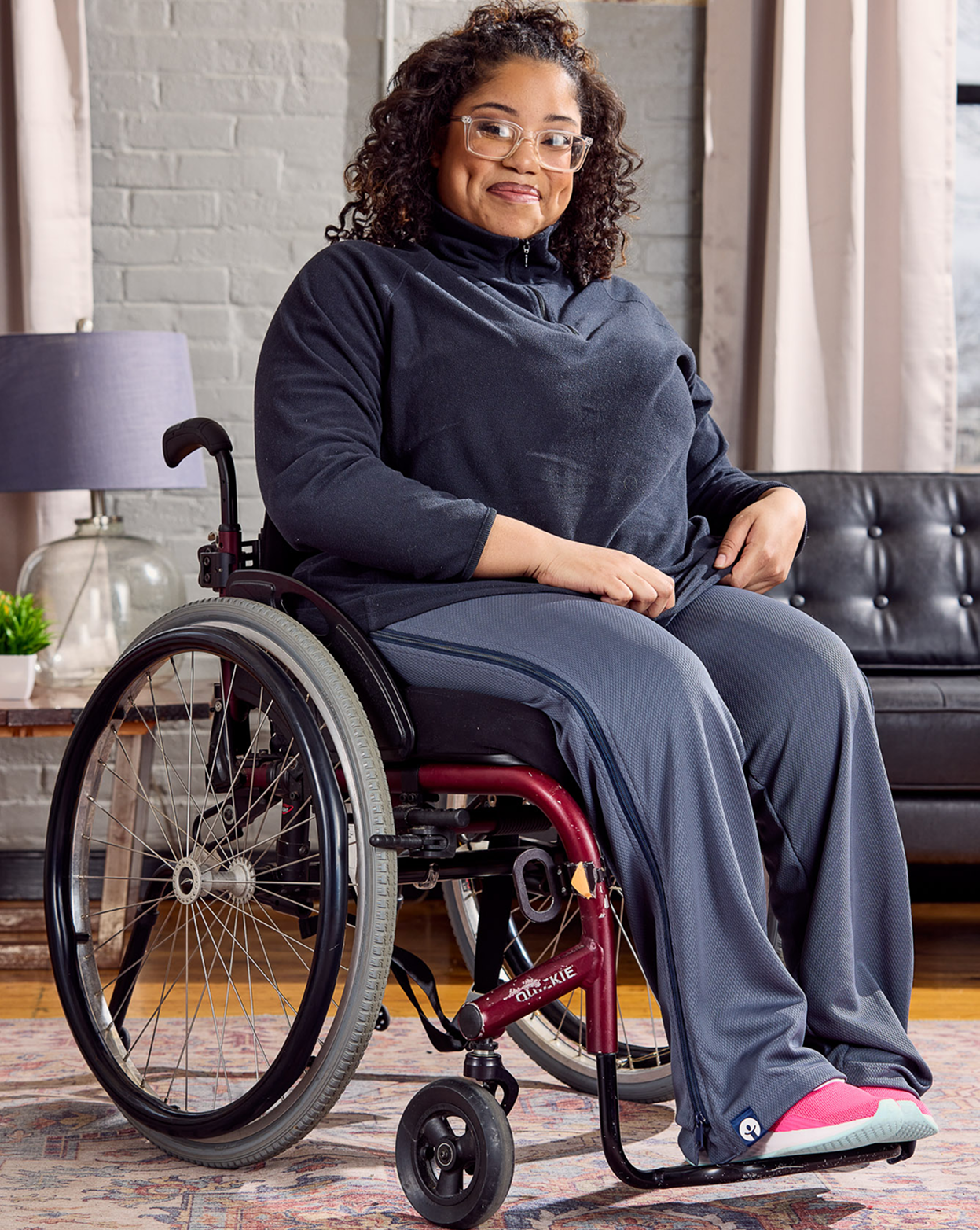 Woman in a wheelchair wearing a dark gray outfit with pink shoes in a living room setting.