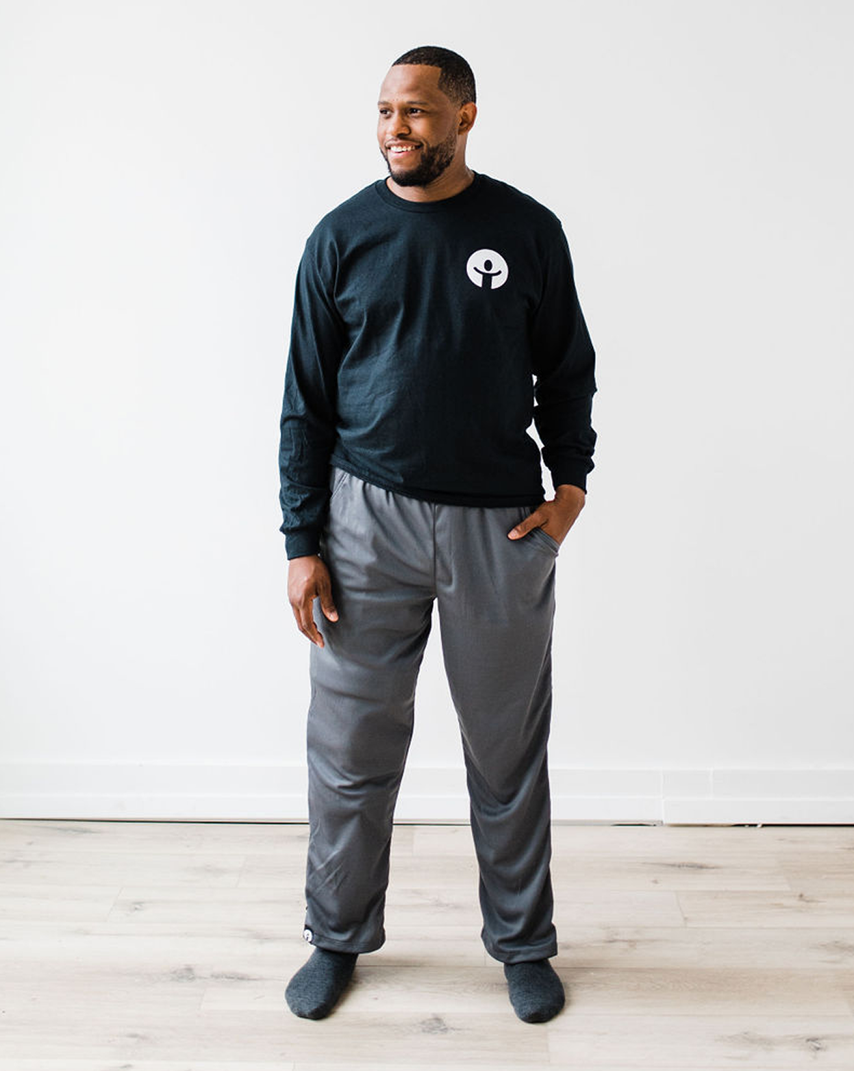 Man wearing a black sweatshirt with a logo and gray pants standing against a white wall.