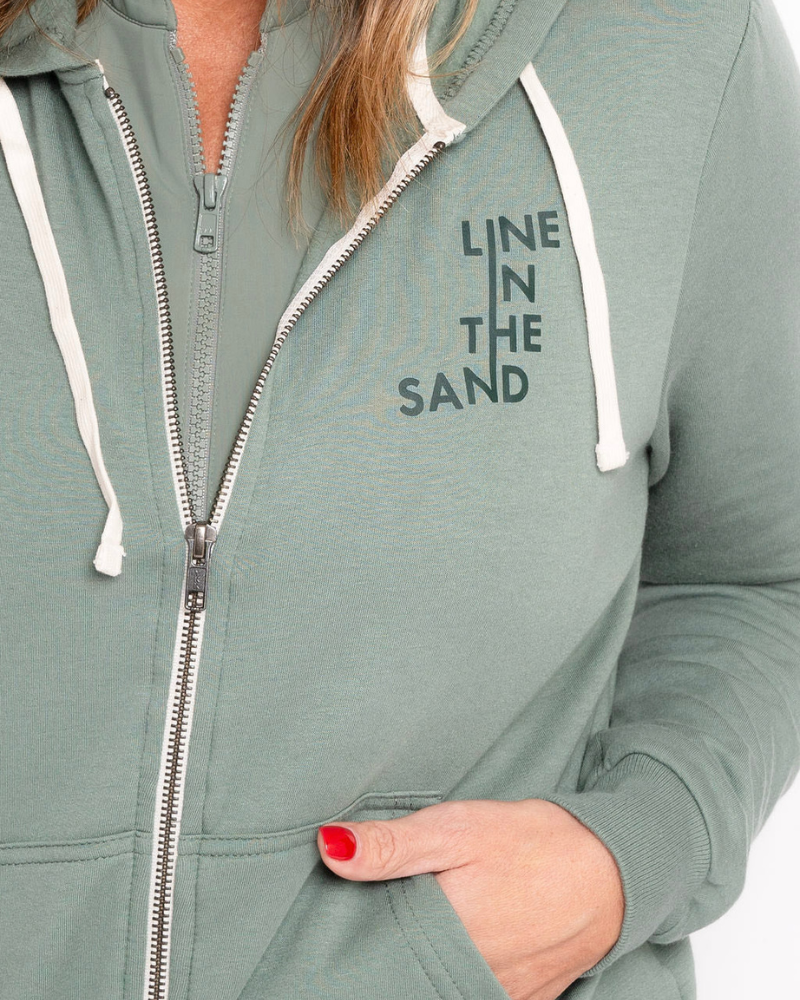 Sage green zip hoodie close-up with front zipper and “LINE IN THE SAND” chest graphic.