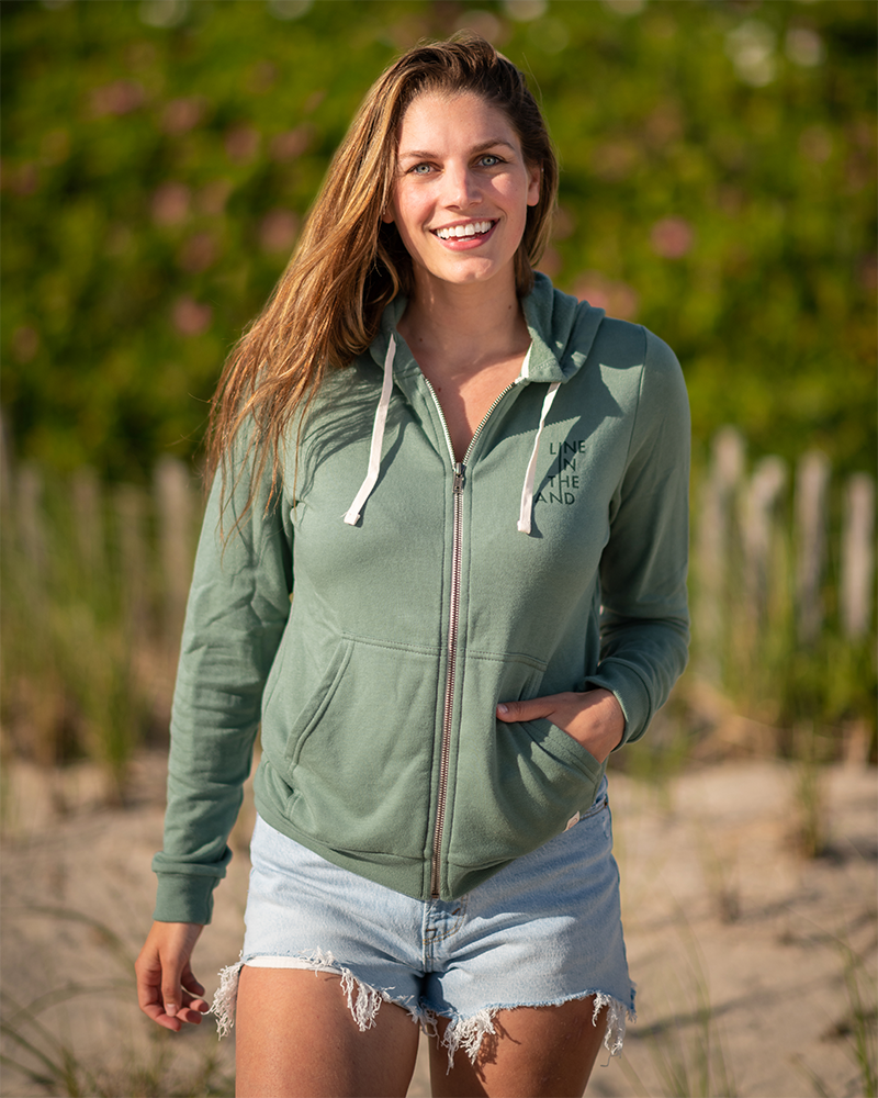 Sage green zip hoodie with white drawstrings and “LINE IN THE SAND” chest graphic.