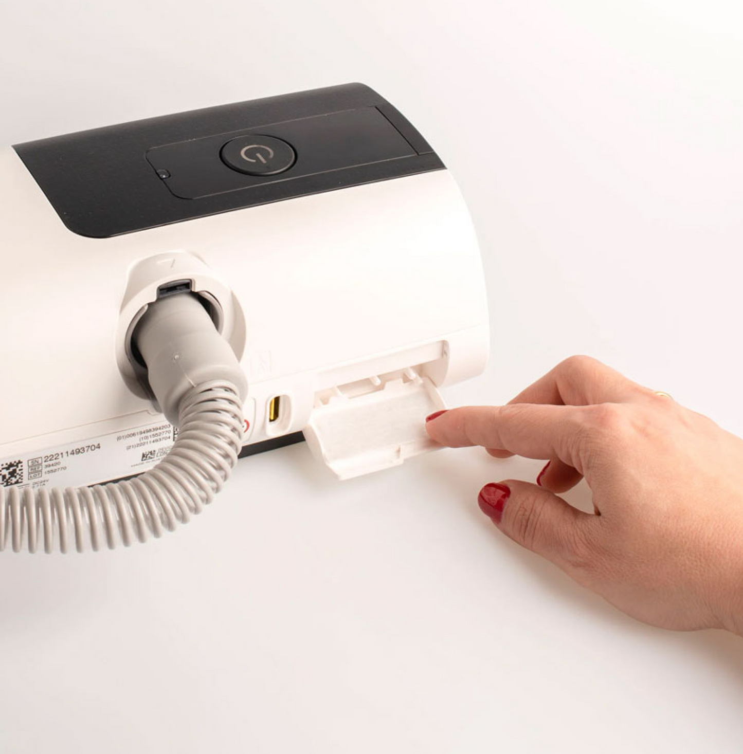 A hand with red nail polish opens a white compartment door to insert an AirSense 11 Filter Pack on a black and white ResMed AirSense 11 device, which features a power button, coiled hose, and attached CPAP filter.