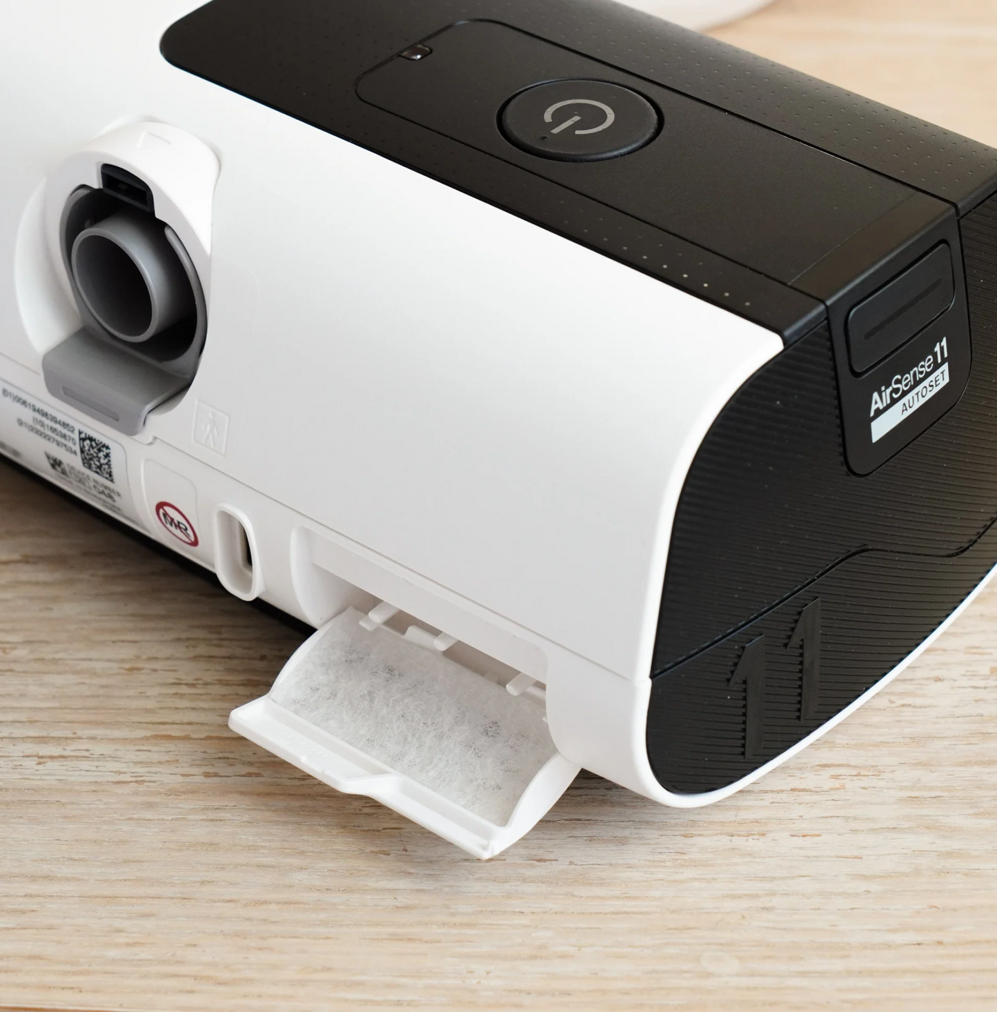 Close-up of AirSense 11 Filter Packs with the CPAP device’s filter tray partially open on a light wooden surface. The power button and vent are visible atop the sleek machine.