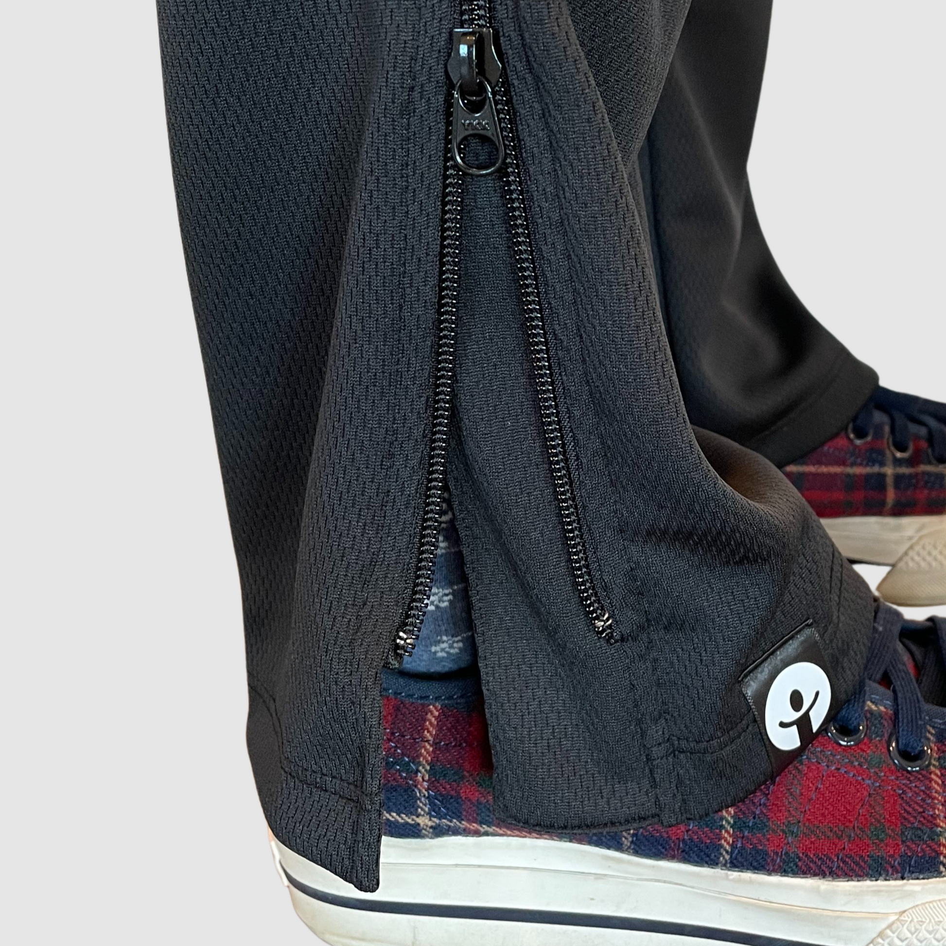 Close-up of a person wearing black pants with a zipper, standing on plaid shoes.