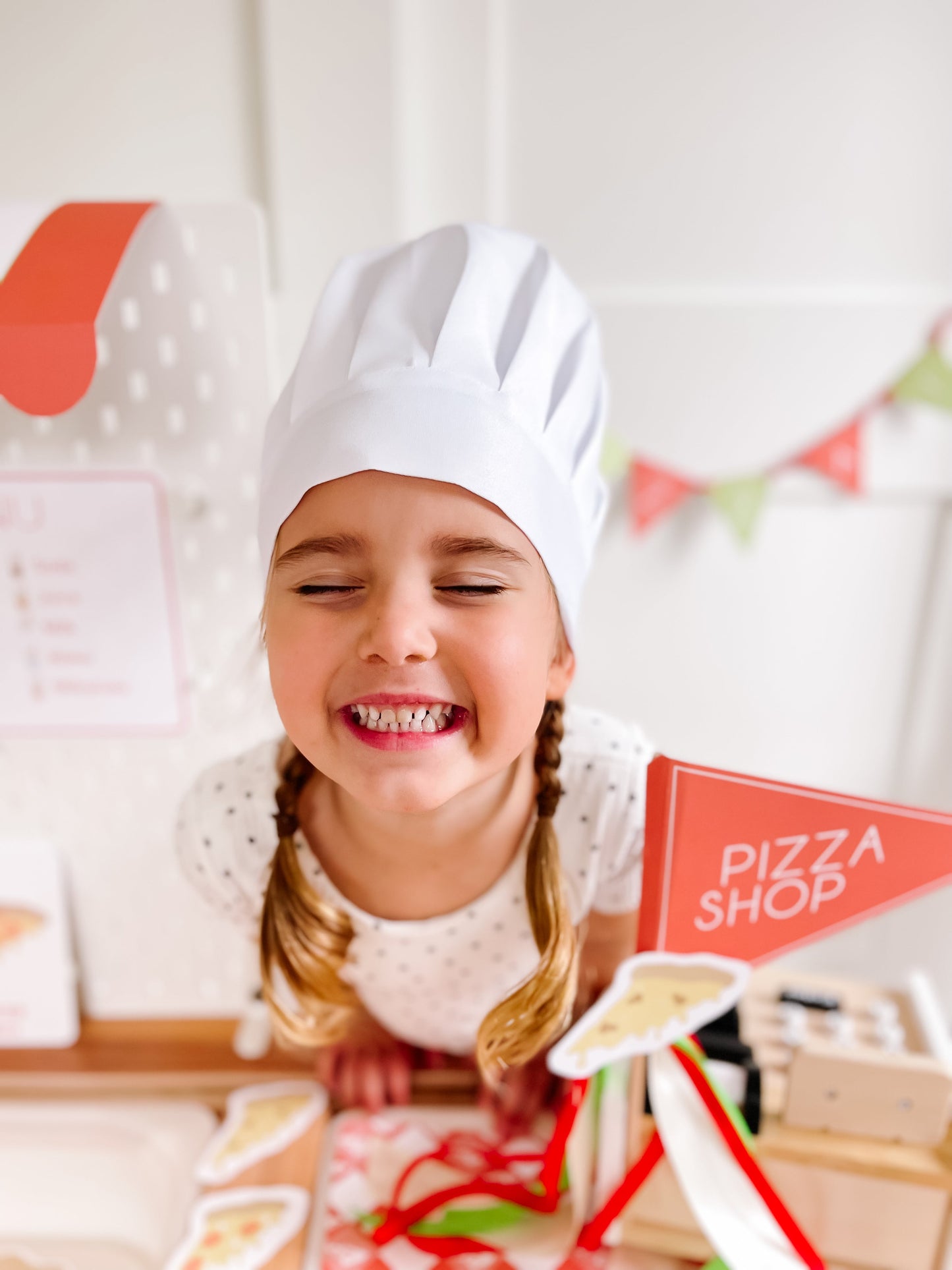 Pizza shop play kit with chef-hat child and red pizza shop sign beside play food