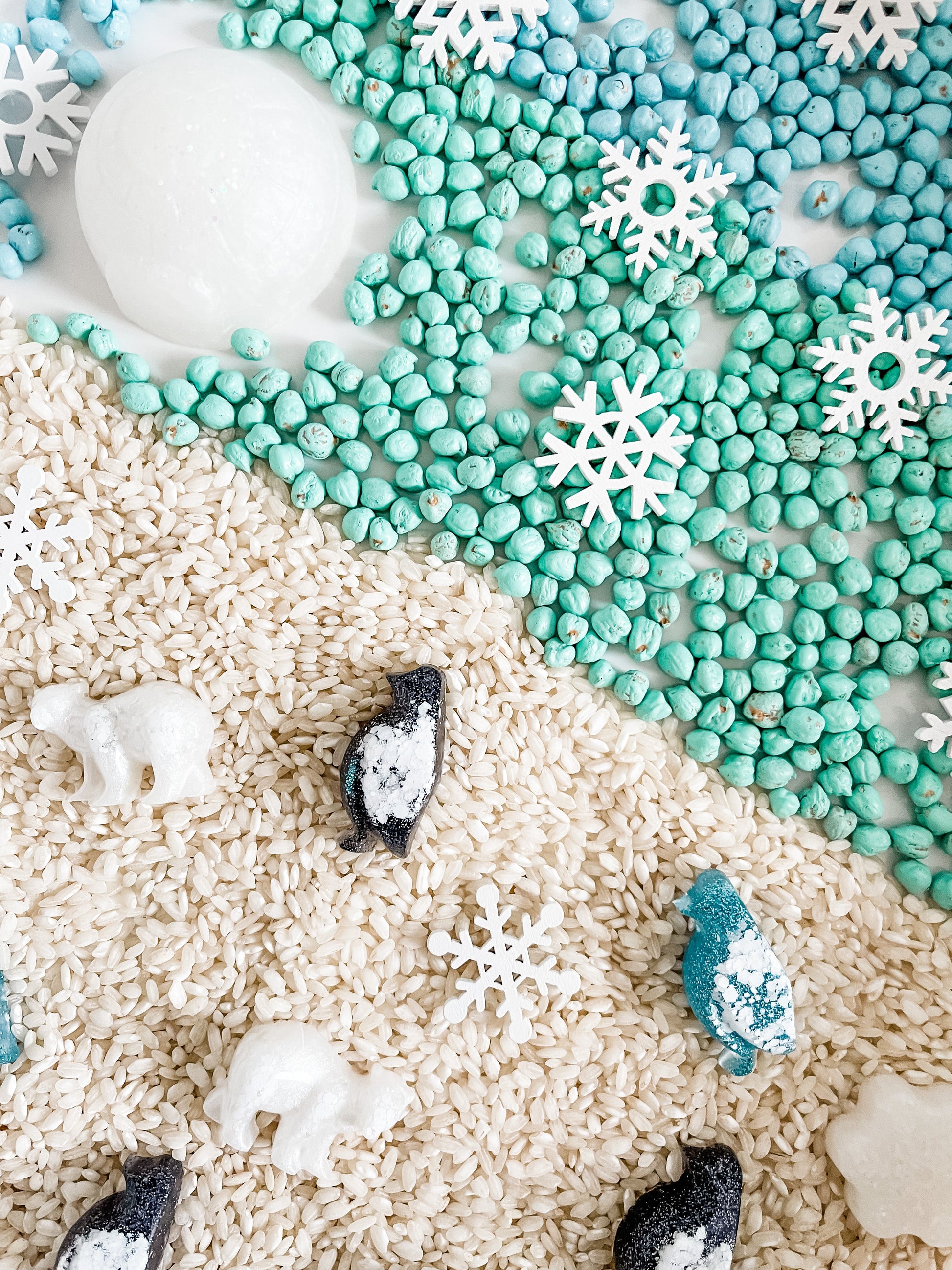 Frosty sensory bin filling with white and teal beads, snowflake bits, and animal pieces