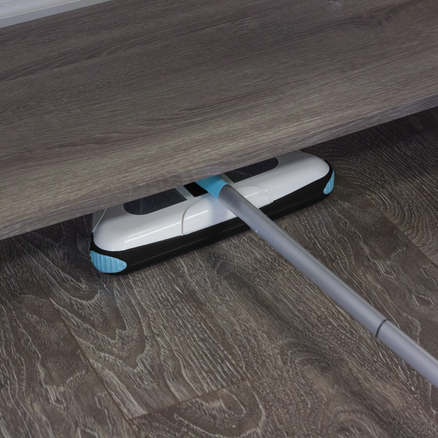 Electric floor sweeper sliding under furniture edge on wood floor, with white and teal parts.