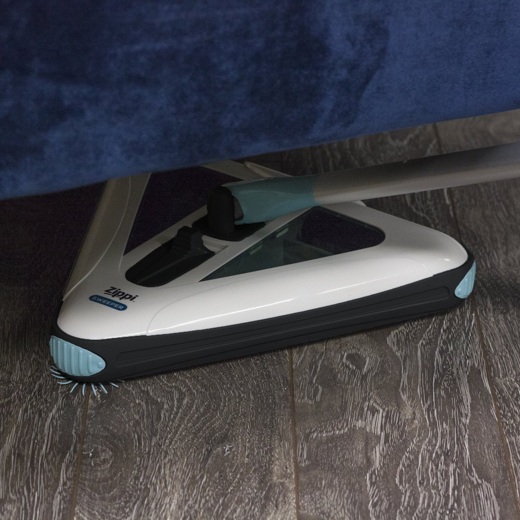 Electric floor sweeper on dark wood floor, with white base and teal side accents.