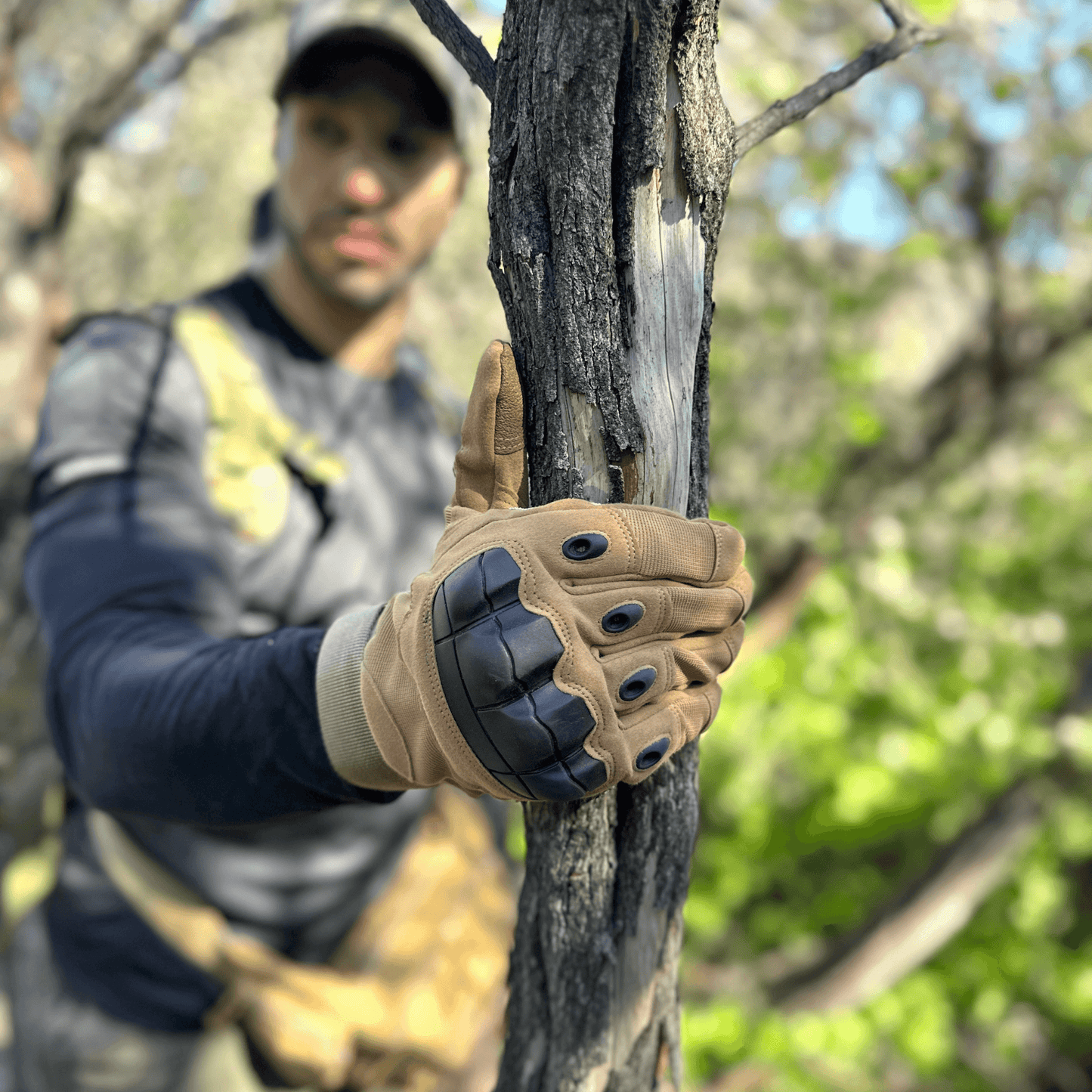 Tan tactical gloves with knuckle pads gripping a tree trunk