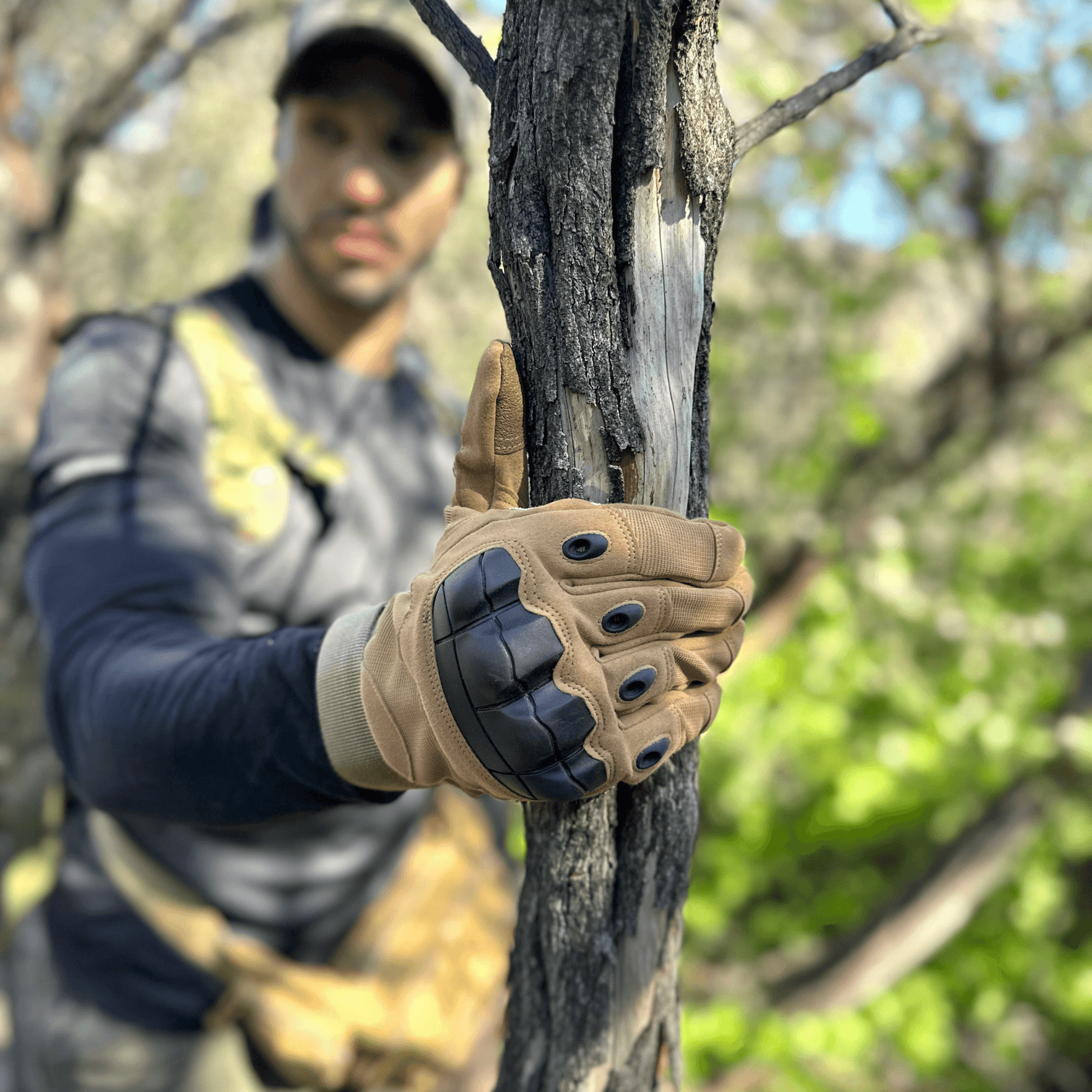 Tan tactical gloves with knuckle pads gripping a tree trunk