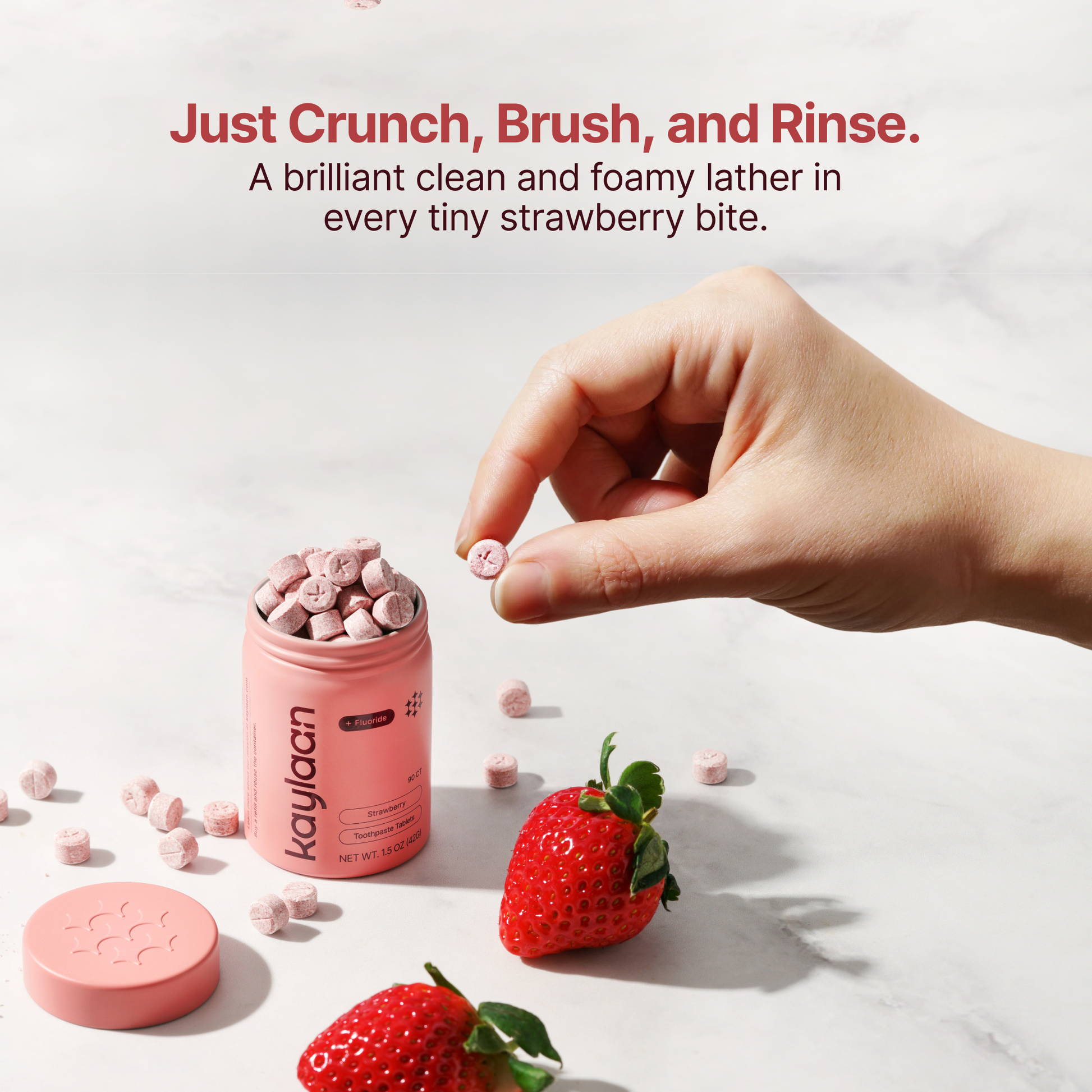 Pink strawberry fluoride toothpaste tablet, with hand near pink container and strawberry