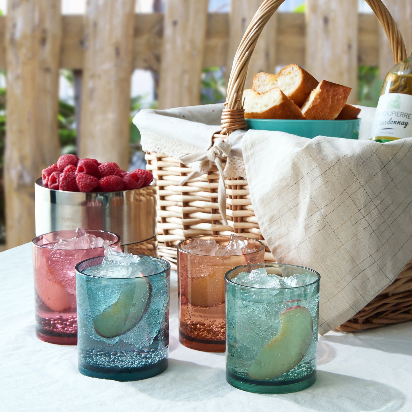 Four acrylic lowball glasses with ice in pink, amber, green, and blue on table