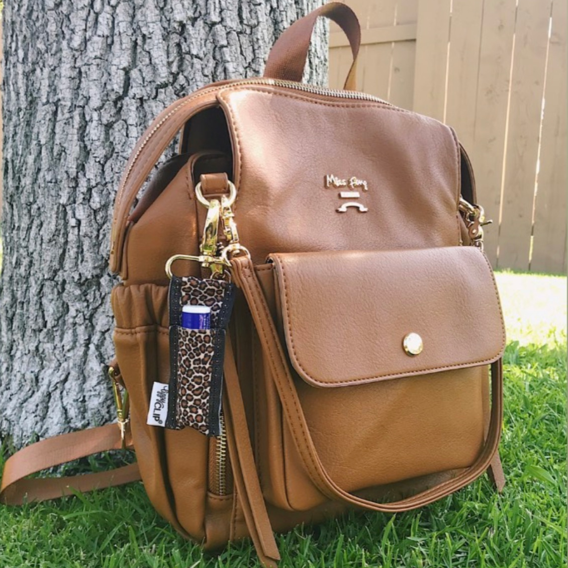 Brown leather backpack with a keychain on grass next to a tree