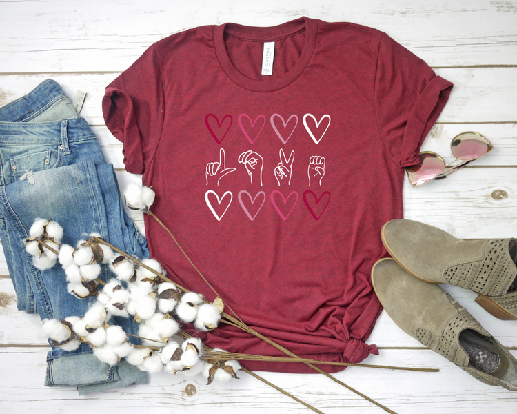 Red t-shirt with heart and hand sign designs, paired with jeans and shoes on a wooden surface.