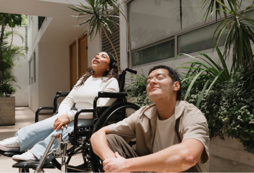 Two people sitting outdoors, one in a wheelchair, with plants and a building in the background.