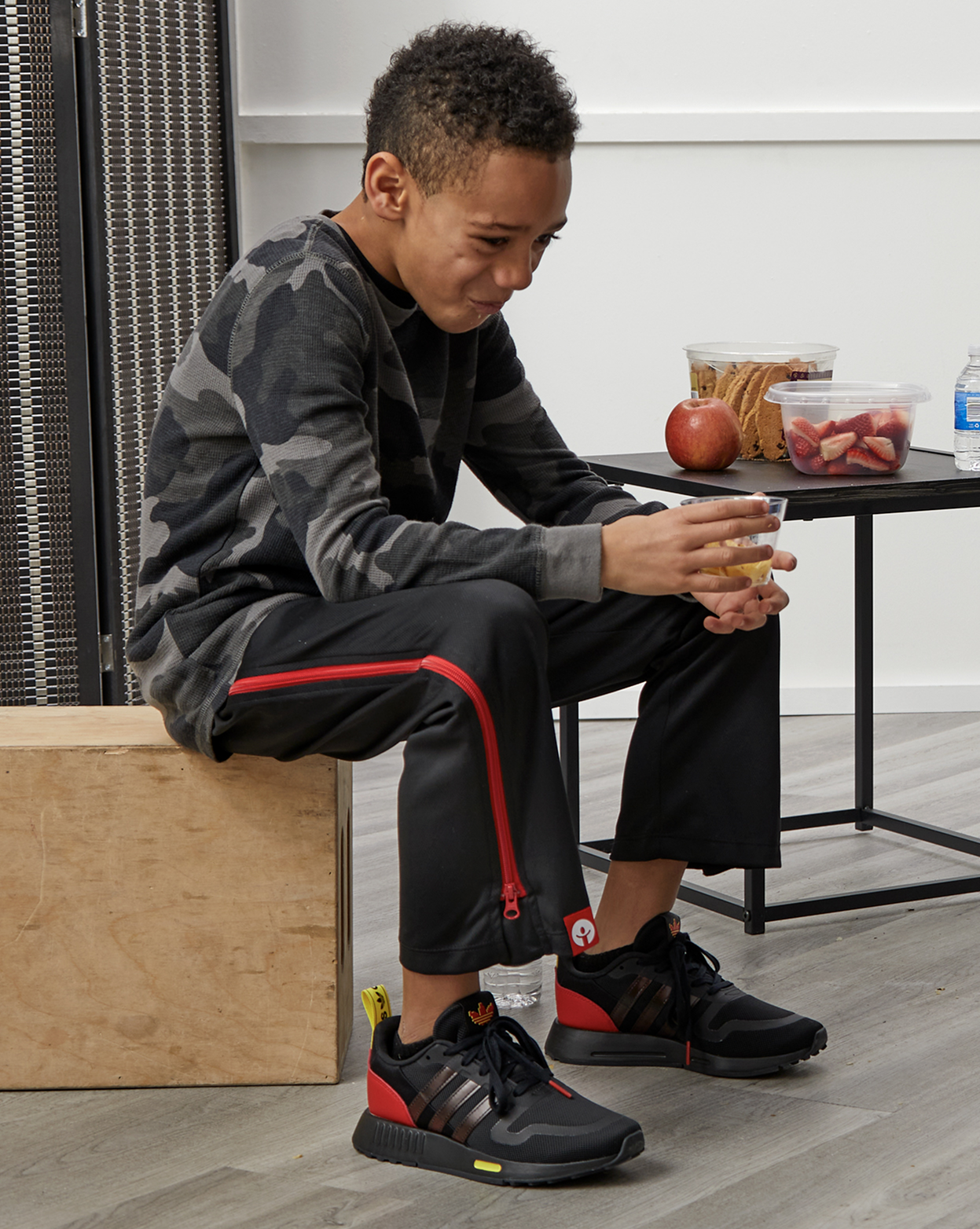 Person sitting on a box wearing black and red athletic shoes, with a table and snacks in the background.
