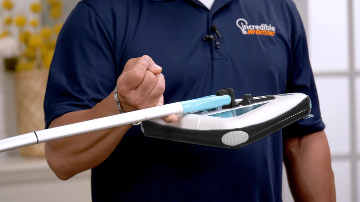 Electric floor sweeper held in hand, with white body and teal handle and cleaning pad.