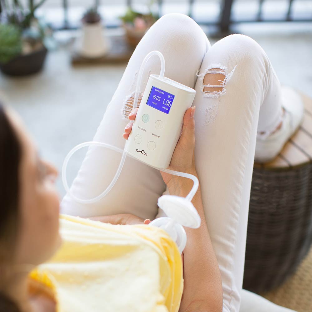 White portable rechargeable breast pump with digital display and clear tubing, by Spectra, held in hand during lifestyle use, side view.