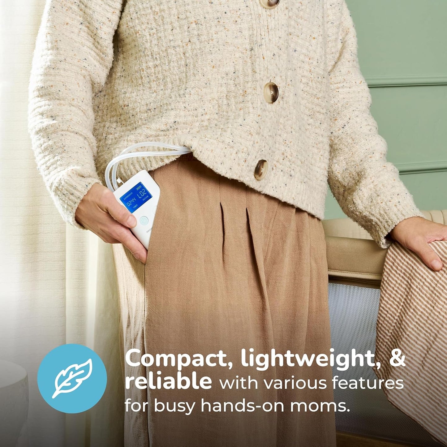 White portable rechargeable breast pump with blue digital display held in hand, worn with beige sweater and brown pants, lifestyle side view