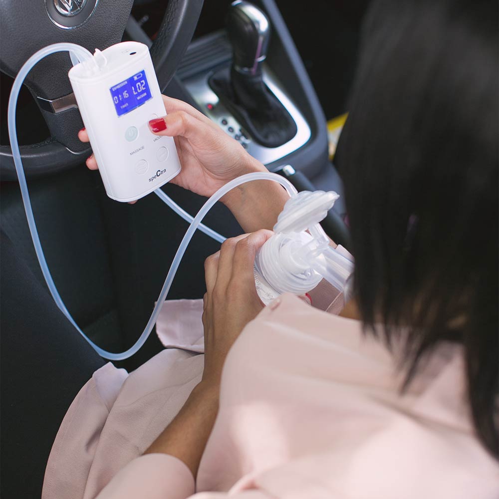 White portable rechargeable breast pump with digital display and clear tubing, by Spectra, shown in use inside a car, held in hand with pink clothing visible.